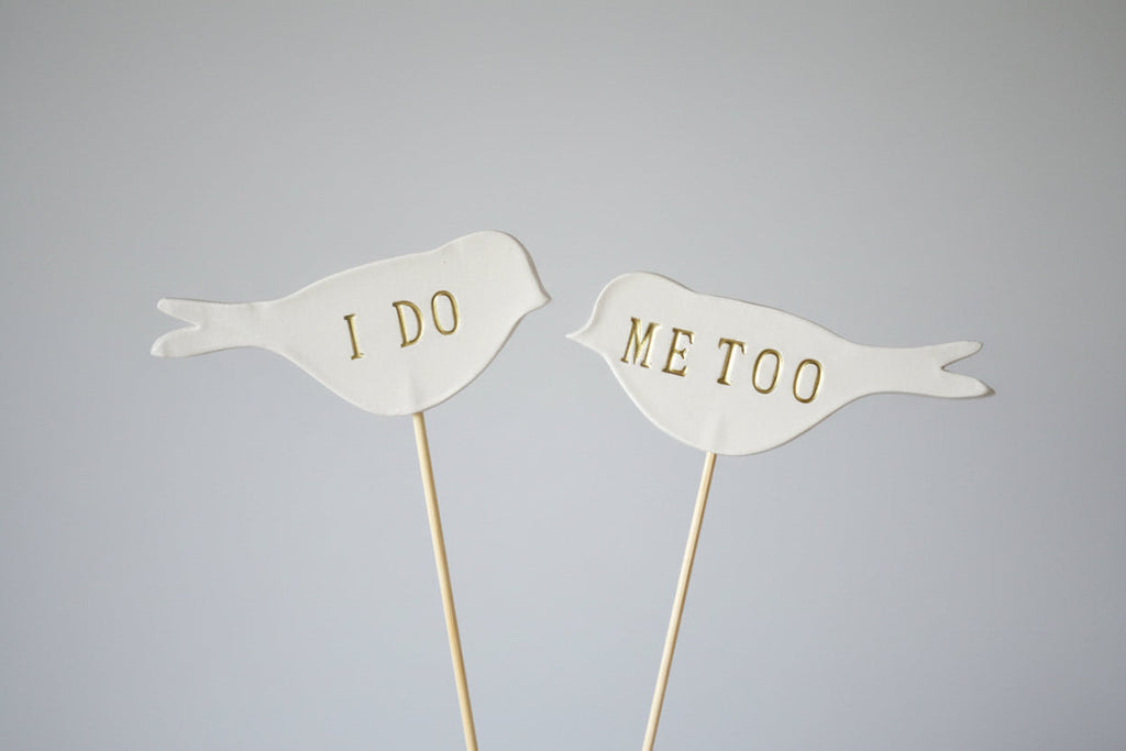 Two white bird-shaped toppers with 'I DO' and 'ME TOO' text on a gray background
