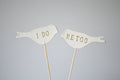 Two white bird-shaped toppers with 'I DO' and 'ME TOO' text on a gray background