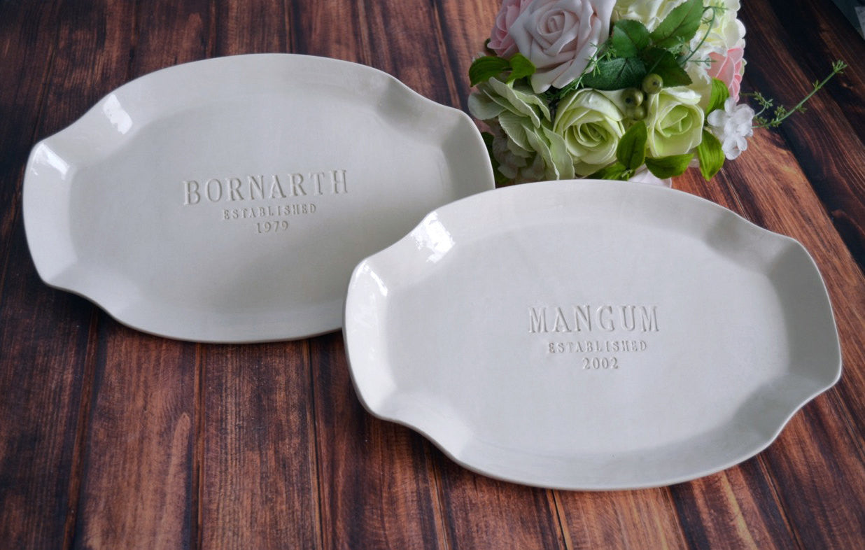 Two white ceramic trays with engraved names on a wooden surface, accompanied by a bouquet of flowers.