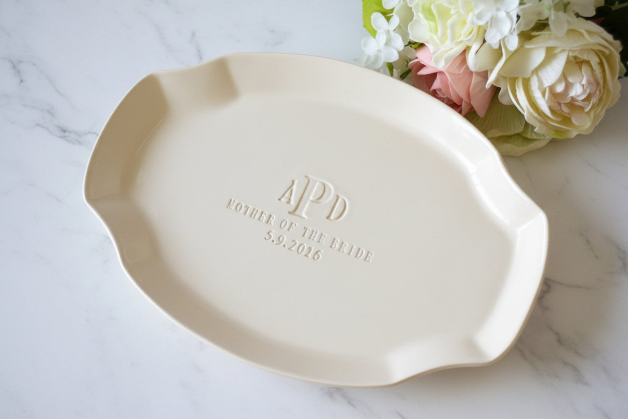 Ivory ceramic platter with 'APD' and 'Bride of the Year 2026' text on a marble surface with flowers.