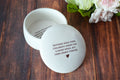 White ceramic trinket dish with engraved text on a wooden surface with flowers in the background
