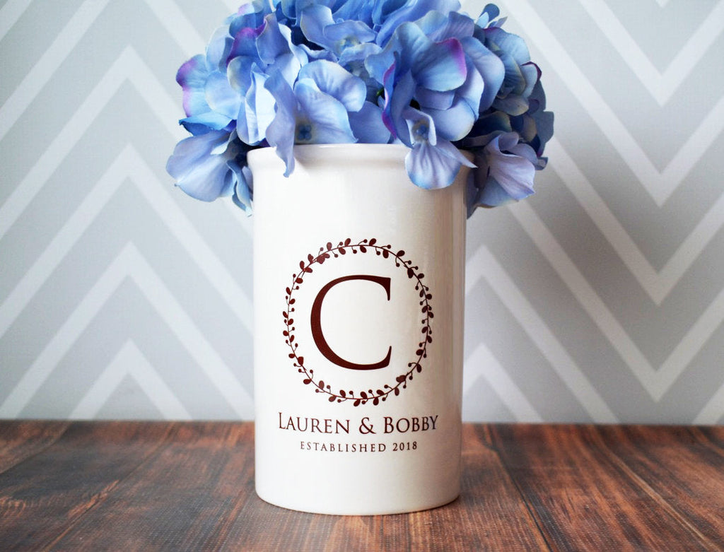 White vase with 'Lauren & Bobby' design and blue flowers on a wooden surface with chevron patterned wall.