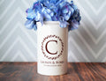 White vase with 'Lauren & Bobby' design and blue flowers on a wooden surface with chevron patterned wall.