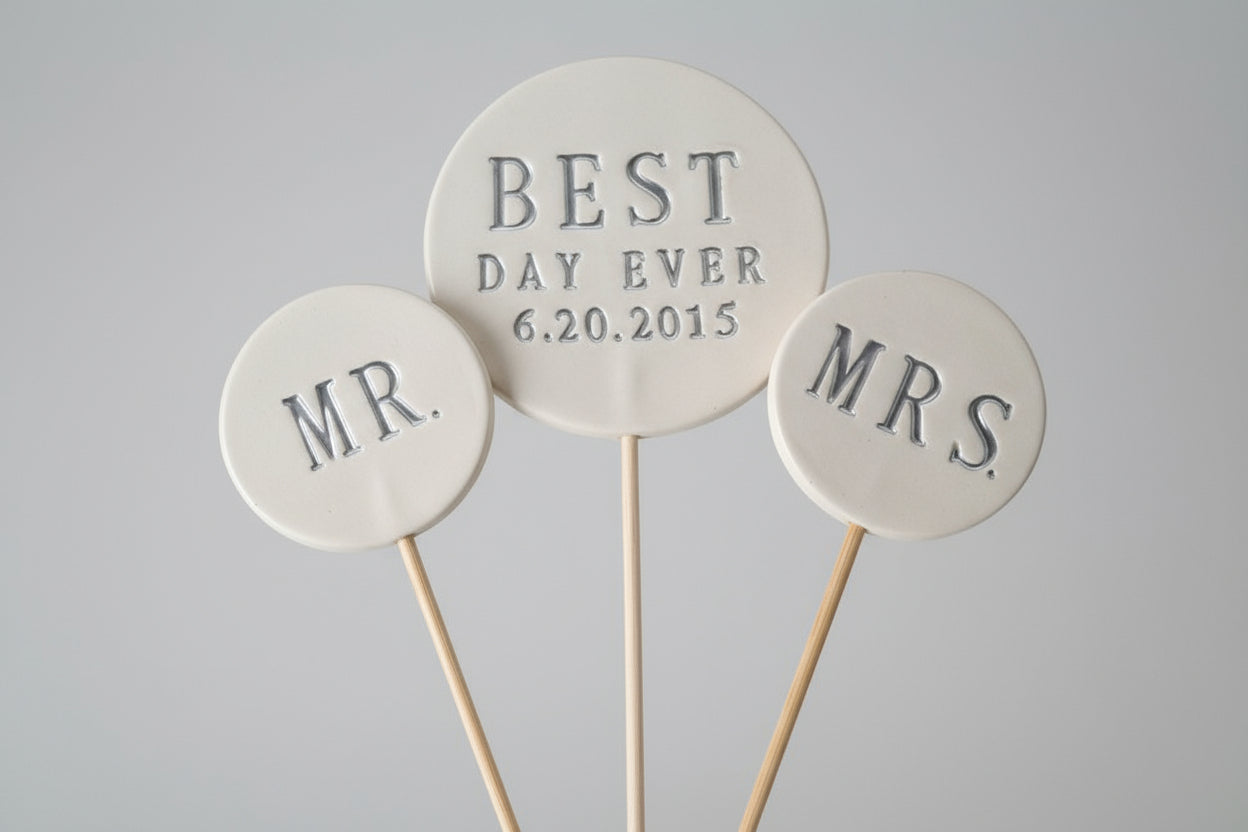 Three round wedding toppers labeled 'MR', 'BEST DAY EVER 6.20.2015', and 'MRS' on a wooden surface.